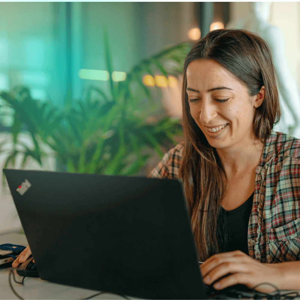 Mulher morena de cabelos longos e blusa xadrez sorri ao receber mensagem de boas-vindas para clientes no notebook